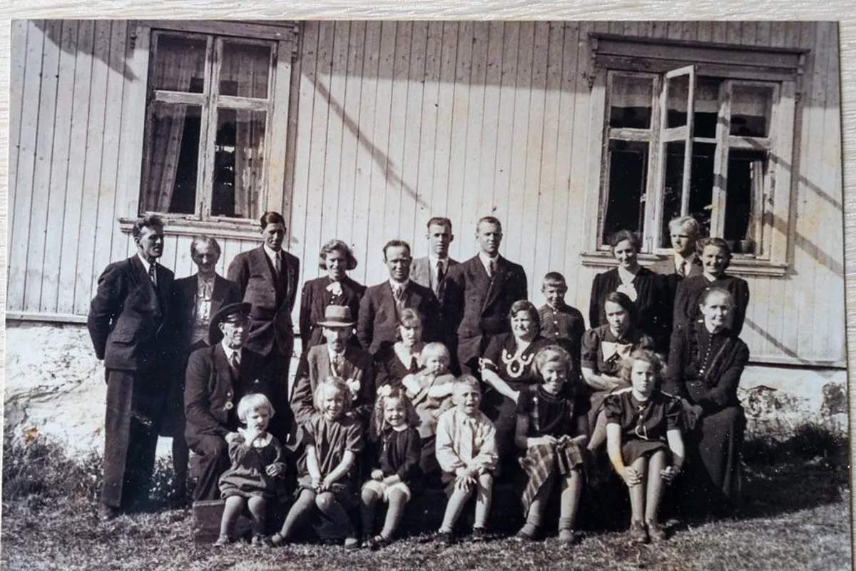 Gammelt gruppebilde av voksne og barn i pent tøy foran et hvitt trehus