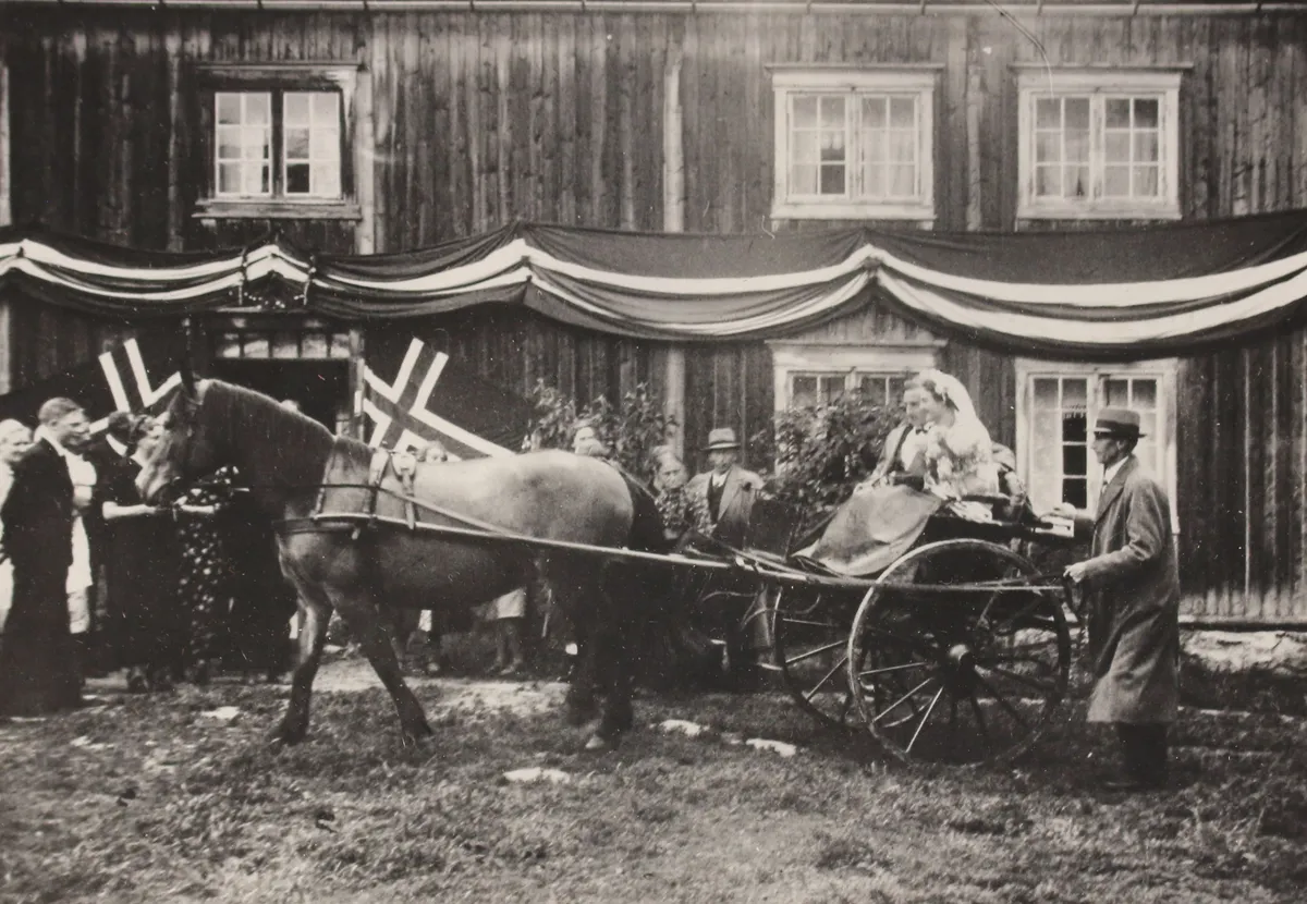 Svart-hvitt-foto: bruden i hest og vogn foran et tømmerhus pyntet med flagg og girlandere, gjester rundt
