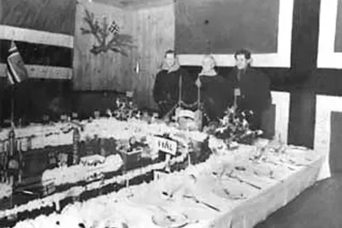 Svart-hvitt-foto av et langbord dekket til festmiddag, med norsk flagg og pynt på veggen