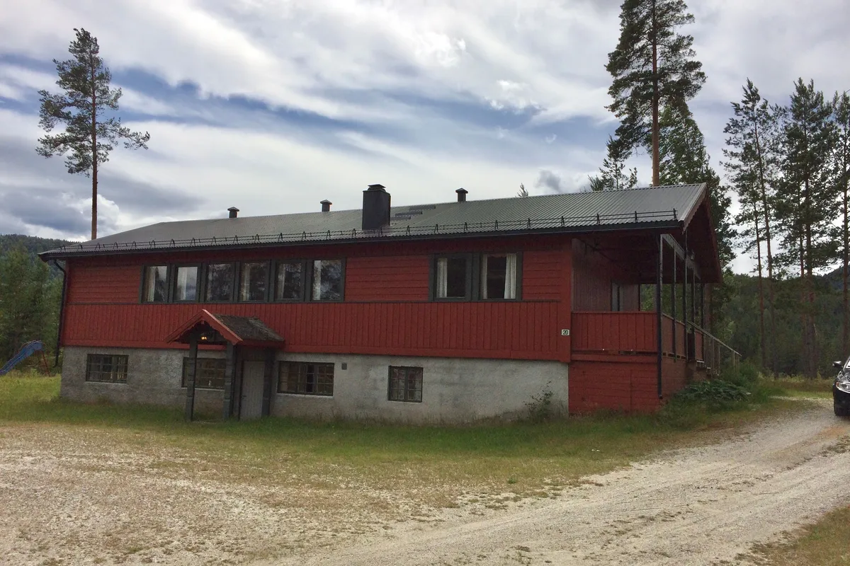 Rødt ungdomshus i to etasjer med grusplass foran og furutrær bak