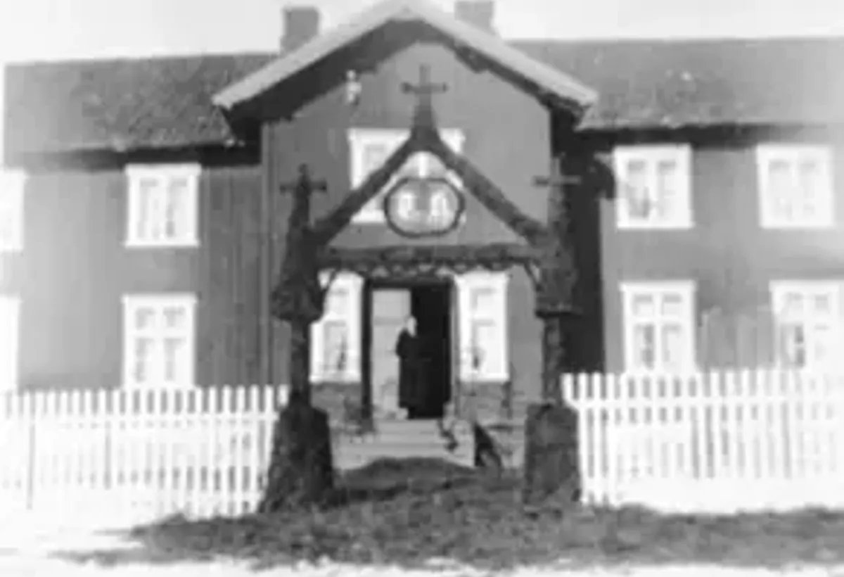 Svart-hvitt-foto av et toetasjes trehus med hvitt stakittgjerde og en portal av granbar over inngangen