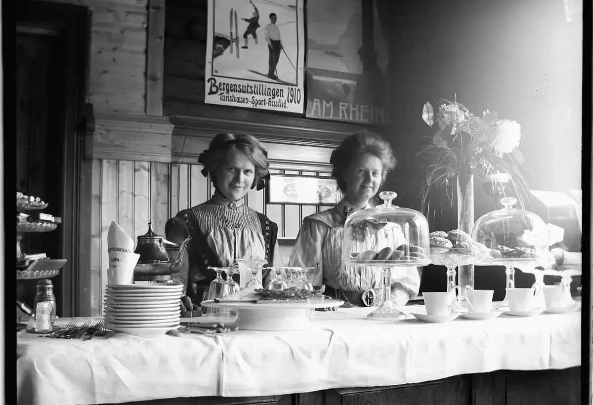 Svart-hvitt-foto av to kvinner bak et dekket kaffebord med kakefat, kopper og blomster