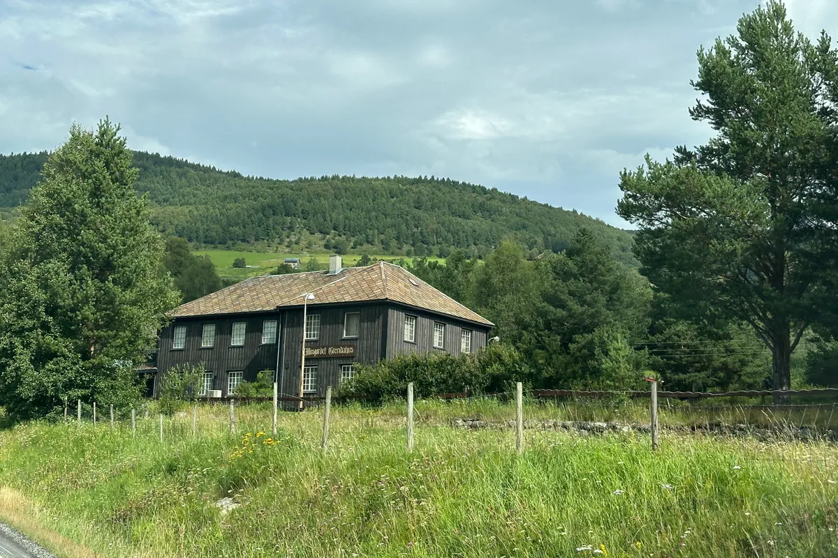Mørkbeiset grendahus i to etasjer med grønn eng foran og skogkledde åser bak