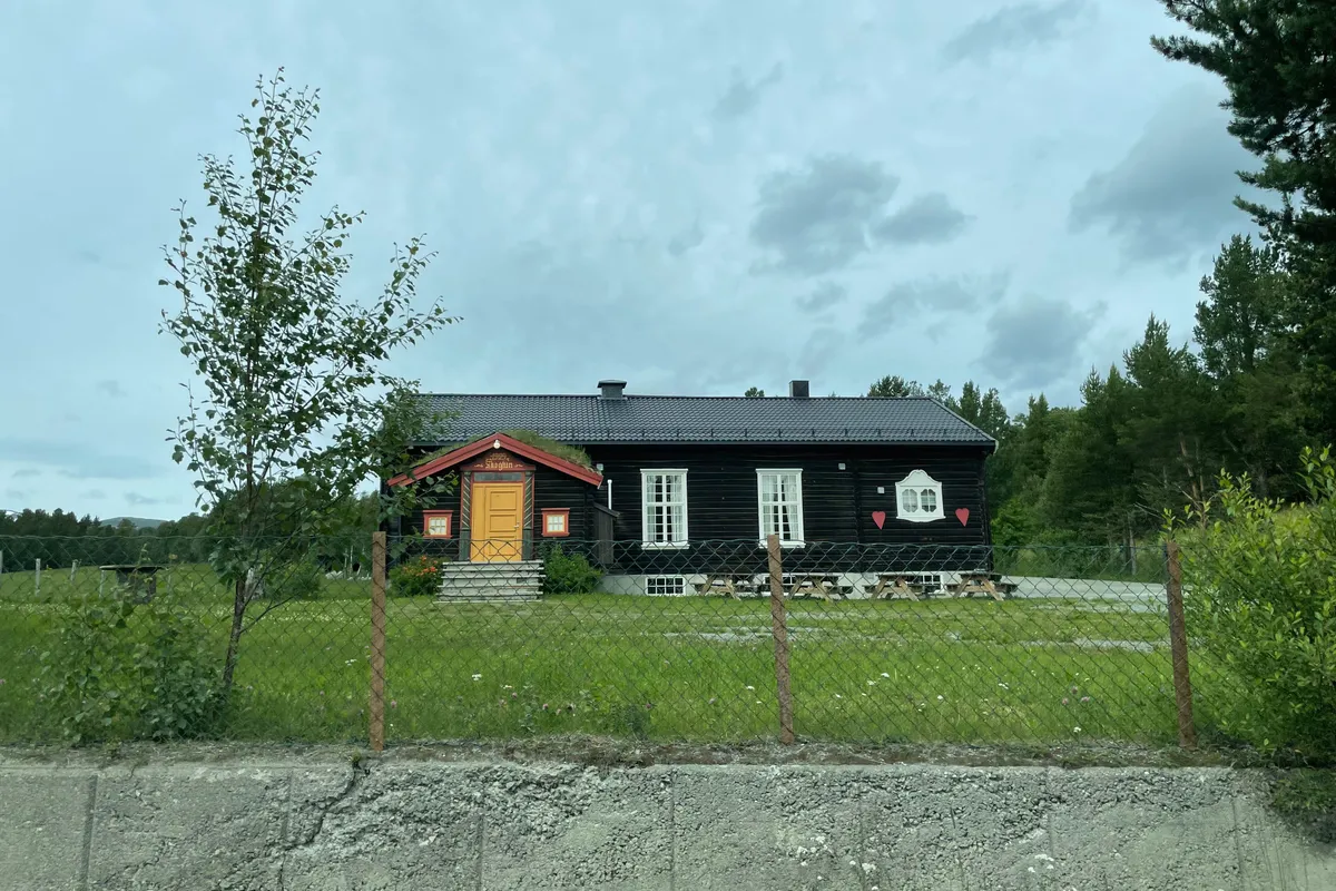 Mørkt ungdomshus med rød dør bak en grønn eng, med skog og åser rundt