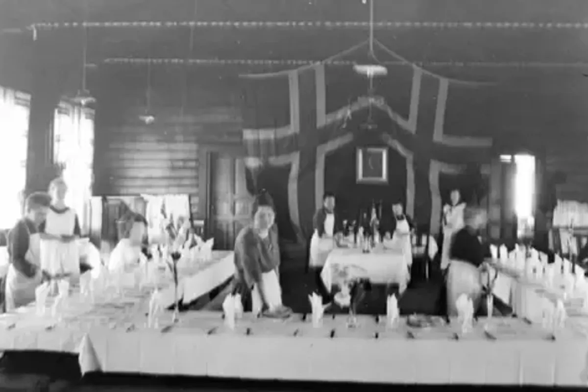 Svart-hvitt-foto av en stor sal pyntet med norske flagg, med dekkede bord og folk som serverer